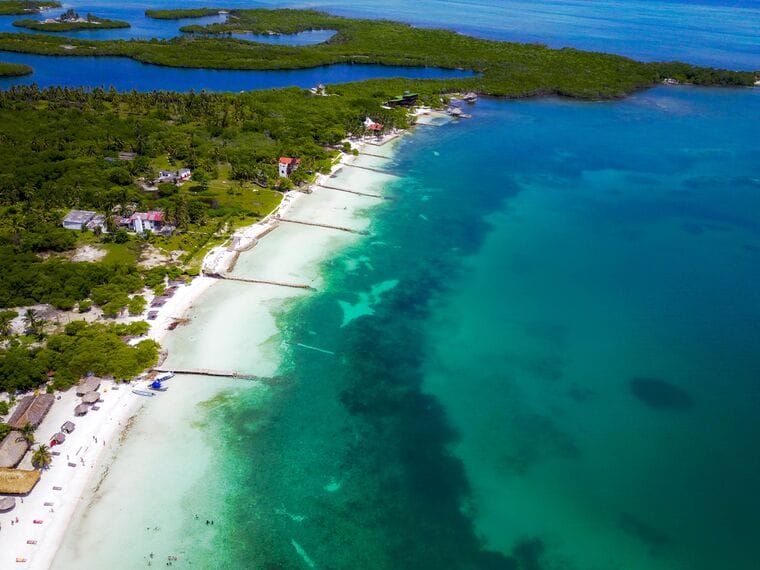 Vista aérea de la zona costera de Cartagena que presenta una playa con aguas cristalinas de color azul verdoso, vegetación adyacente y edificios dispersos. Varios muelles se adentran en el agua y a lo lejos se ven pequeñas islas como la Isla Tintipán. Perfecto para una escapada de 3 días.