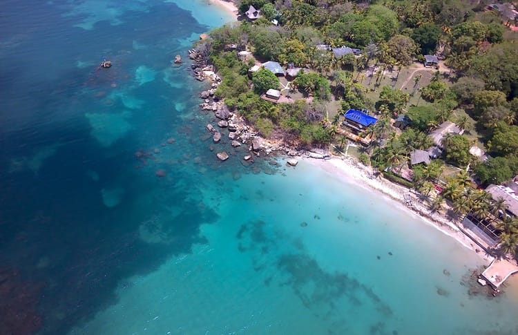 Vista aérea del litoral de San Bernardo del Viento con aguas cristalinas de color turquesa, playa de arena blanca, zonas rocosas y vegetación verde. Edificios dispersos salpican la porción terrestre a lo largo de la costa.