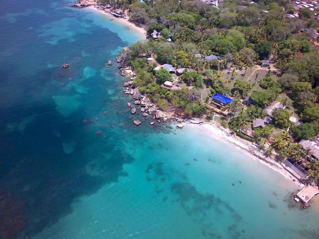 Vista aérea de una costa tropical en San Bernardo del Viento con aguas cristalinas de color turquesa, costa rocosa y exuberante vegetación. Varios edificios y cabañas se encuentran esparcidos entre los árboles cerca de la playa.