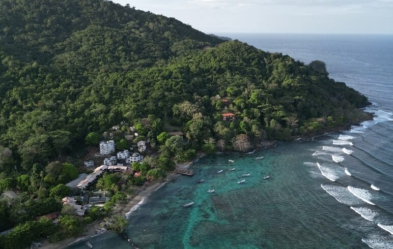 Vista aérea de una ladera costera verde y exuberante en Capurganá, con edificios enclavados, bordeada por un océano turquesa con barcos en el agua y olas acercándose a la orilla, un escenario perfecto para tu próximo plan de viaje para descubrir Necoclí.