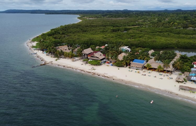 Vista aérea de la playa tropical de Cartagena con edificios con techo de paja, palmeras y un denso bosque verde al fondo. Las tranquilas aguas del océano azul verdoso rodean la costa arenosa de Rincón del Mar, perfecta para una escapada tranquila de 3 días.