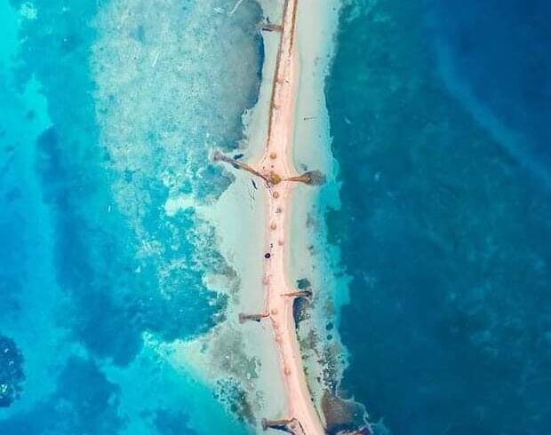 Vista aérea de un puente estrecho que conecta un pequeño edificio con tierra en Isla Palma, rodeado de agua azul clara y formaciones de coral visibles, perfecto para una escapada de 3 días desde Montería.