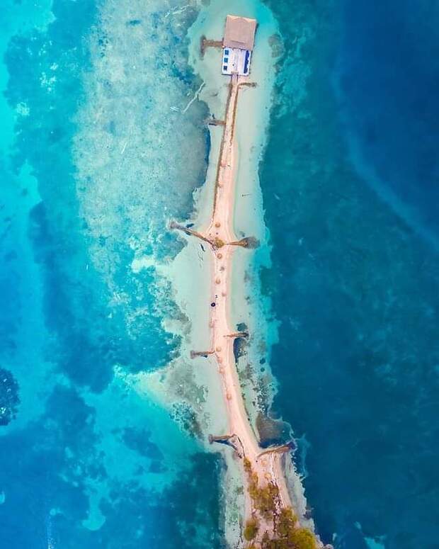 Vista aérea de un puente estrecho que conecta un pequeño edificio con tierra en Isla Palma, rodeado de agua azul clara y formaciones de coral visibles, perfecto para una escapada de 3 días desde Montería.