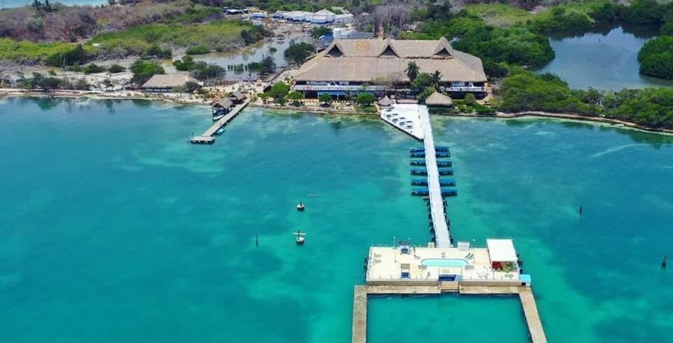 Vista aérea de un resort en una isla tropical, Isla Palma, con un muelle de madera que se extiende hacia aguas turquesas, rodeado de exuberante vegetación y bordeado por el océano. Esta joya escondida cerca de Cartagena ofrece un escape perfecto con su serena porción terrestre que abraza la belleza de la naturaleza.