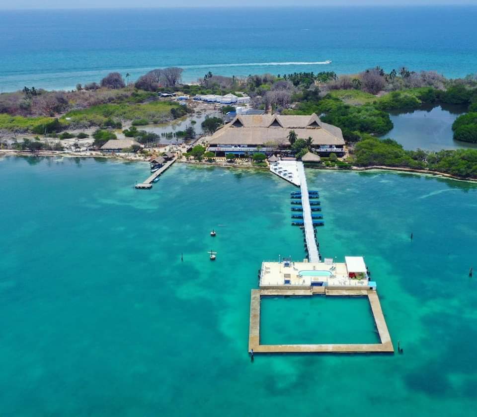 Vista aérea de un resort en una isla tropical, Isla Palma, con un muelle de madera que se extiende hacia aguas turquesas, rodeado de exuberante vegetación y bordeado por el océano. Esta joya escondida cerca de Cartagena ofrece un escape perfecto con su serena porción terrestre que abraza la belleza de la naturaleza.