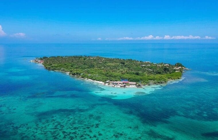 Vista aérea de Isla Múcura, una pequeña isla tropical cerca de Cartagena, rodeada de aguas cristalinas y arrecifes de coral. La isla presenta una exuberante vegetación verde y edificios dispersos visibles desde arriba. Perfecto para una escapada de 3 días.