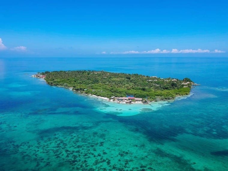 Vista aérea de Isla Múcura, una pequeña isla tropical cerca de Cartagena, rodeada de aguas cristalinas y arrecifes de coral. La isla presenta una exuberante vegetación verde y edificios dispersos visibles desde arriba. Perfecto para una escapada de 3 días.