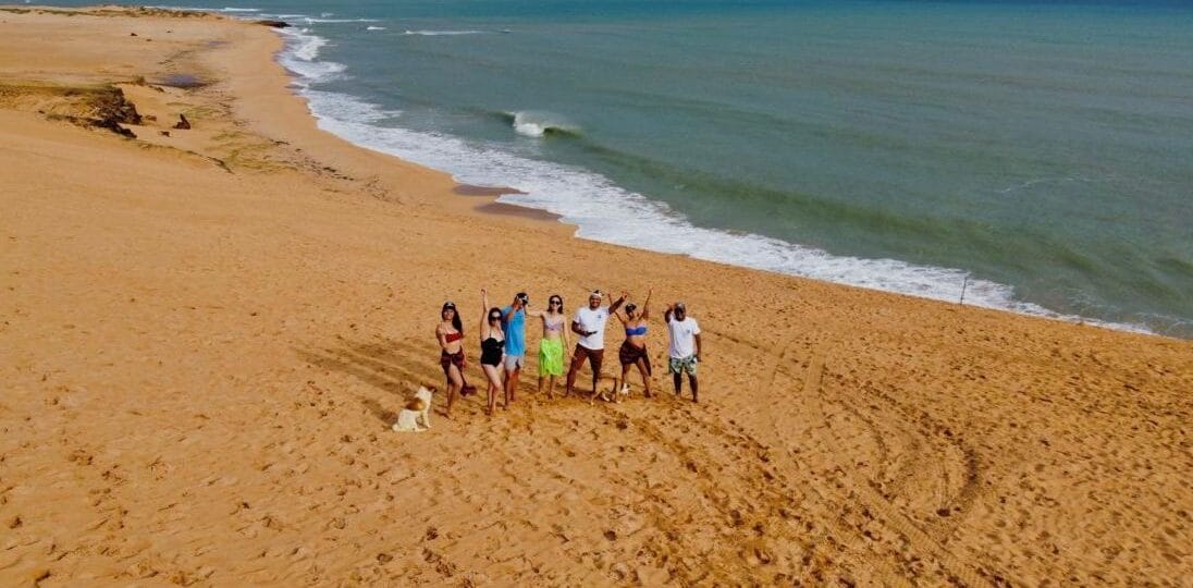 Un grupo de personas se encuentran juntas en una playa de arena cerca del océano en la Alta Guajira, con un perro cerca. El cielo está parcialmente nublado y se ven olas a lo lejos.