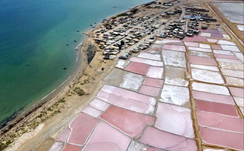 Vista aérea de salinas de color rosa cerca de un pequeño pueblo costero en la Alta Guajira, adyacente a un mar verde y una playa de arena, a solo 5 días de viaje desde Riohacha.