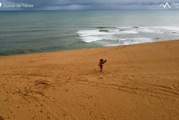 Una persona se encuentra sola en las vastas dunas de arena de la Alta Guajira, cerca del océano en Dunas de Taroa, con olas rompiendo al fondo bajo un cielo nublado. Esta impresionante escena es sólo un punto culminante de un inolvidable viaje de 5 días desde Riohacha.