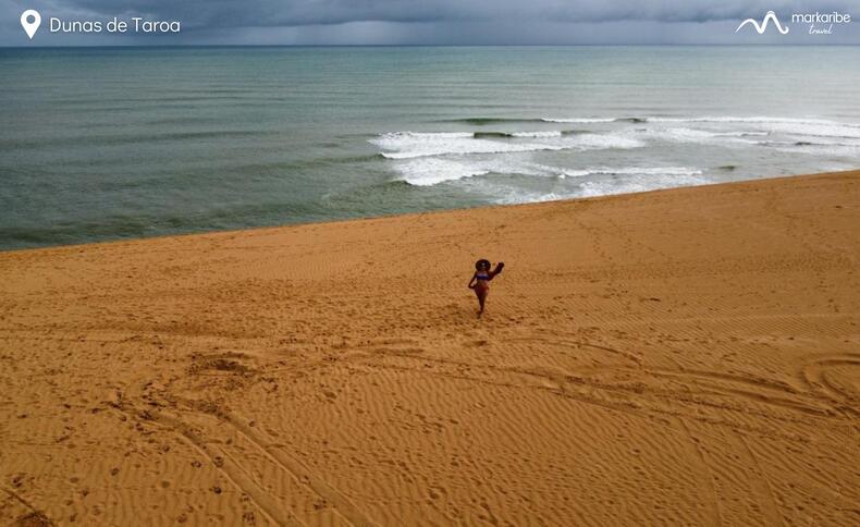 Una persona se encuentra sola en las vastas dunas de arena de la Alta Guajira, cerca del océano en Dunas de Taroa, con olas rompiendo al fondo bajo un cielo nublado. Esta impresionante escena es sólo un punto culminante de un inolvidable viaje de 5 días desde Riohacha.