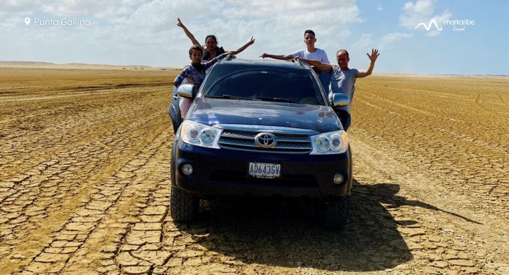 Un grupo de personas posan sobre y al lado de una camioneta azul estacionada en un paisaje desértico seco y agrietado bajo un cielo parcialmente nublado en Punta Gallina, Alta Guajira.