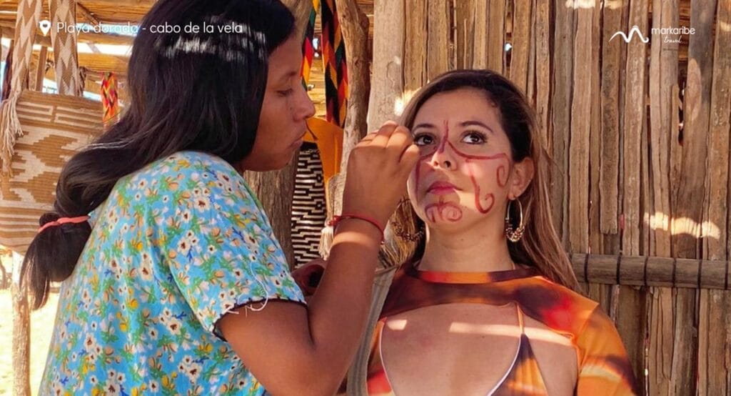 Una persona está pintando diseños intrincados en el rostro de una mujer. Se encuentran en un entorno rústico con estructuras de madera al fondo. El texto de la imagen dice "Playa Arguello - cabo de la vela, Riohacha". Experimente el arte y la belleza de la Alta Guajira durante 5 días.