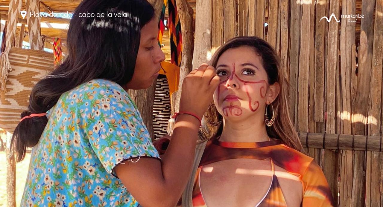 Una persona está pintando diseños intrincados en el rostro de una mujer. Se encuentran en un entorno rústico con estructuras de madera al fondo. El texto de la imagen dice "Playa Arguello - cabo de la vela, Riohacha". Experimente el arte y la belleza de la Alta Guajira durante 5 días.