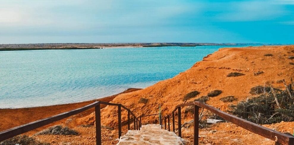 Unos escalones de piedra con barandillas de madera conducen a una serena masa de agua azul, rodeada de un terreno árido y rocoso bajo un cielo despejado, que recuerda al Cabo de la Vela. Esta impresionante vista ofrece un escape tranquilo para quienes viajan durante 4 días a través de esta hermosa porción terrestre.