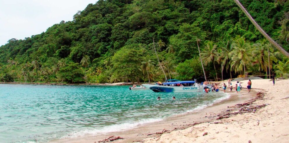 La gente disfruta de una playa tropical en Necoclí con aguas cristalinas, exuberante vegetación y barcos anclados cerca de la orilla.