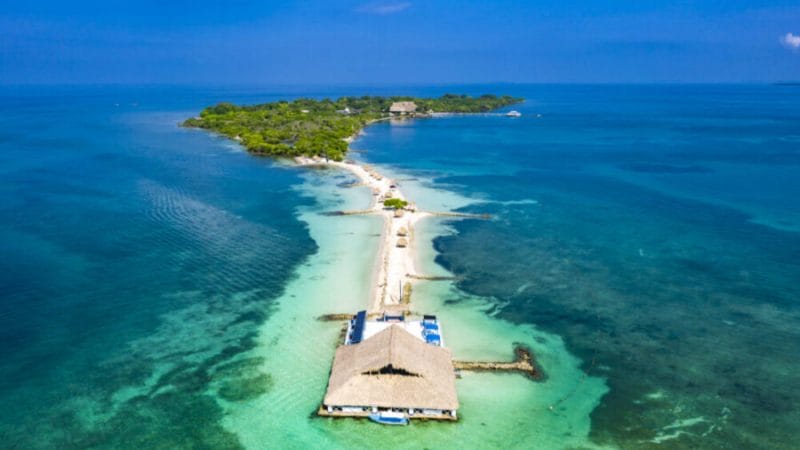 Vista aérea de Isla Palma, una isla tropical con exuberante vegetación, un camino de arena que conduce a un edificio y aguas turquesas circundantes.