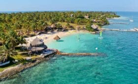 Un resort tropical en Isla Múcura, con palmeras, cabañas y una playa de arena se extiende hacia el océano turquesa. Se pueden ver barcos y muelles cerca de la costa, lo que lo convierte en la escapada perfecta para 3 días de relajación después de explorar Cartagena.