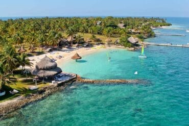 Un resort tropical en Isla Múcura, con palmeras, cabañas y una playa de arena se extiende hacia el océano turquesa. Se pueden ver barcos y muelles cerca de la costa, lo que lo convierte en la escapada perfecta para 3 días de relajación después de explorar Cartagena.