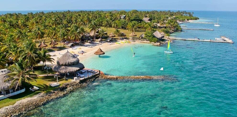 Un resort tropical en Isla Múcura, con palmeras, cabañas y una playa de arena se extiende hacia el océano turquesa. Se pueden ver barcos y muelles cerca de la costa, lo que lo convierte en la escapada perfecta para 3 días de relajación después de explorar Cartagena.