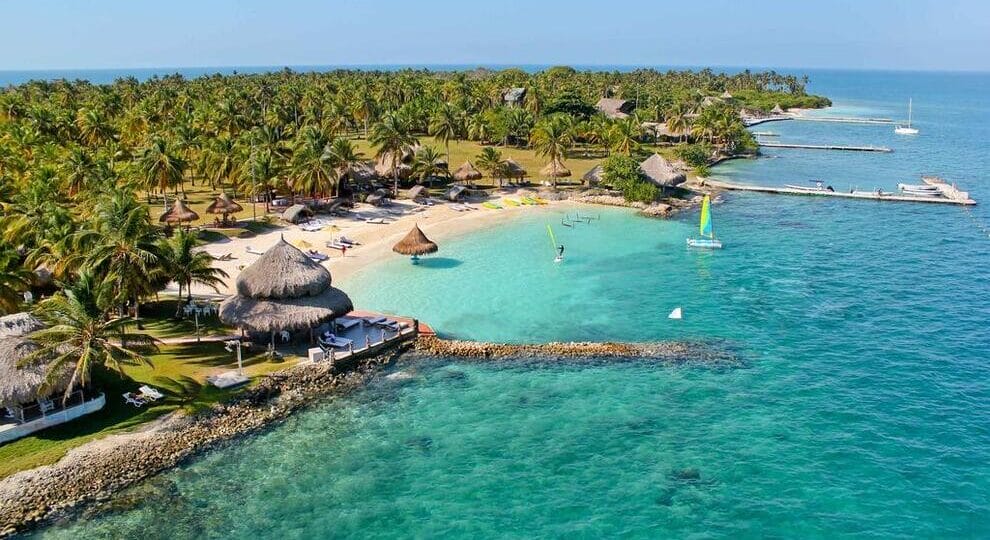 Un resort tropical en Isla Múcura, con palmeras, cabañas y una playa de arena se extiende hacia el océano turquesa. Se pueden ver barcos y muelles cerca de la costa, lo que lo convierte en la escapada perfecta para 3 días de relajación después de explorar Cartagena.