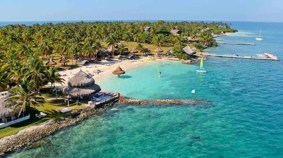 Explora las Islas Secretas del Caribe Colombiano: Los Paraísos Mejor Guardados Un resort tropical en Isla Múcura, con palmeras, cabañas y una playa de arena se extiende hacia el océano turquesa. Se pueden ver barcos y muelles cerca de la costa, lo que lo convierte en la escapada perfecta para 3 días de relajación después de explorar Cartagena.