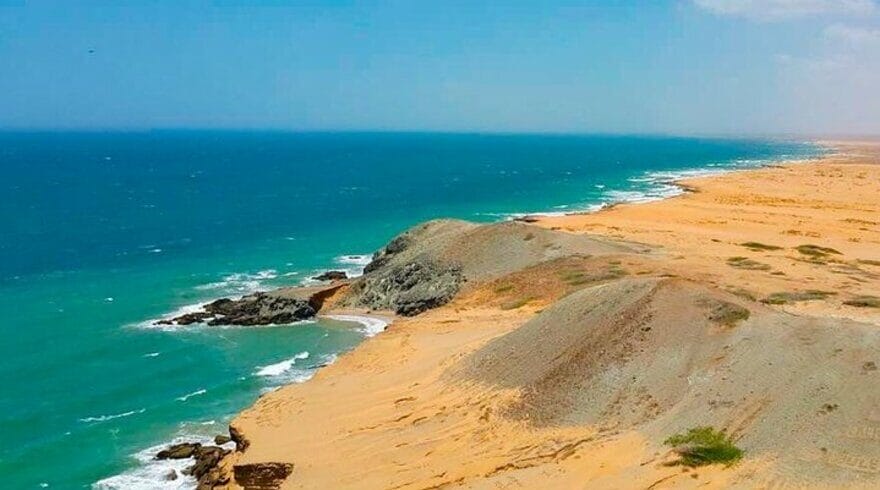 Un paisaje costero en Riohacha, Alta Guajira, presenta una vasta playa de arena con suaves colinas que se encuentran con el océano turquesa bajo un cielo parcialmente nublado. En las dunas se ve escasa vegetación.