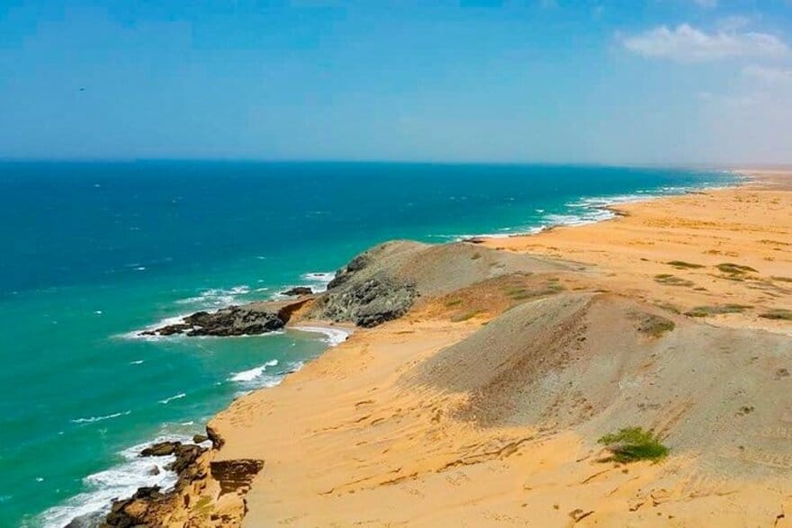 Un paisaje costero en Riohacha, Alta Guajira, presenta una vasta playa de arena con suaves colinas que se encuentran con el océano turquesa bajo un cielo parcialmente nublado. En las dunas se ve escasa vegetación.