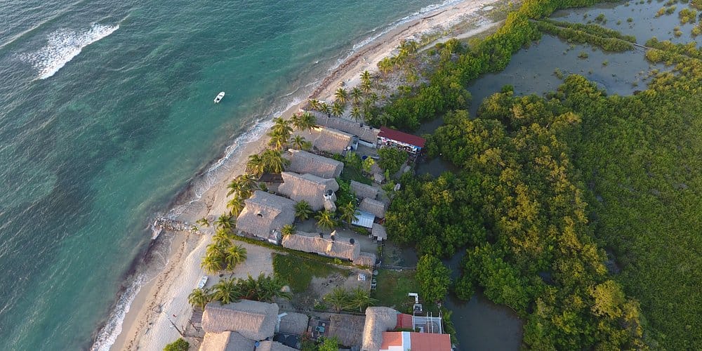 Vista aérea de cabañas frente a la playa en Rincón del Mar, rodeadas de palmeras y adyacentes a un exuberante y verde bosque de manglares con un pequeño bote en la costa cercana, en las afueras de Cartagena.