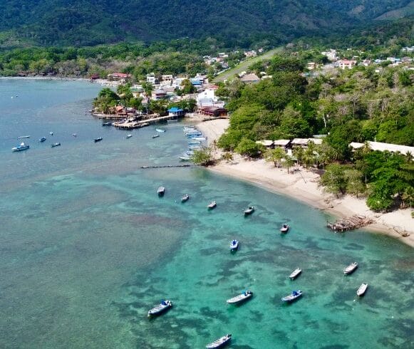 Vista aérea de un pueblo costero con barcos anclados en aguas cristalinas de color turquesa cerca de una playa de arena, rodeado de exuberantes colinas verdes.