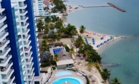 Vista aérea del complejo costero con altos edificios azules, una piscina con un patrón único y una playa con sombrillas junto a aguas tranquilas.