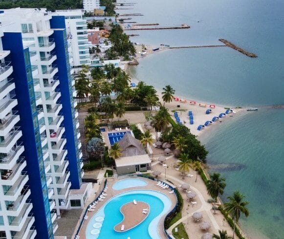 Vista aérea del complejo costero con altos edificios azules, una piscina con un patrón único y una playa con sombrillas junto a aguas tranquilas.