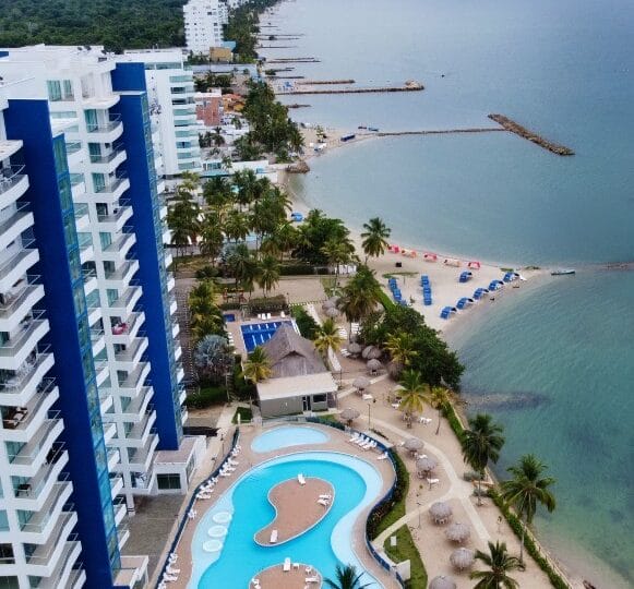 Vista aérea del complejo costero con altos edificios azules, una piscina con un patrón único y una playa con sombrillas junto a aguas tranquilas.