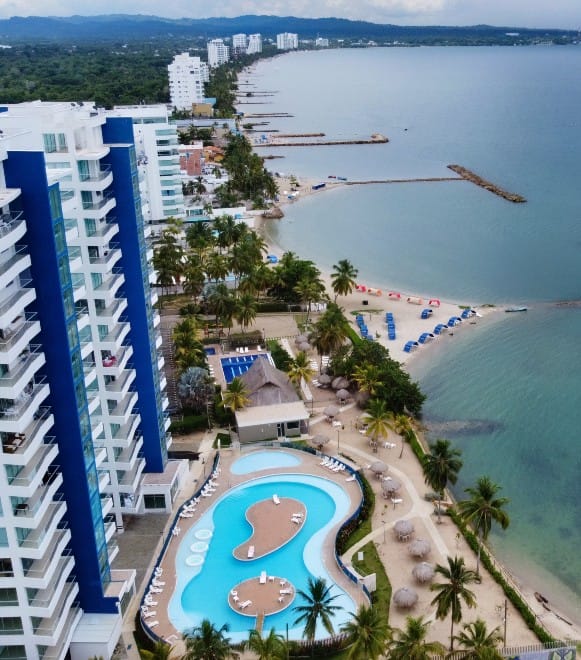 Vista aérea del complejo costero con altos edificios azules, una piscina con un patrón único y una playa con sombrillas junto a aguas tranquilas.