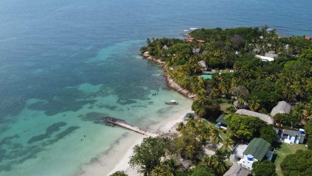 Vista aérea de una costa tropical con aguas cristalinas de color turquesa, una pequeña playa de arena, un barco atracado y una densa vegetación que se extiende hasta la costa.