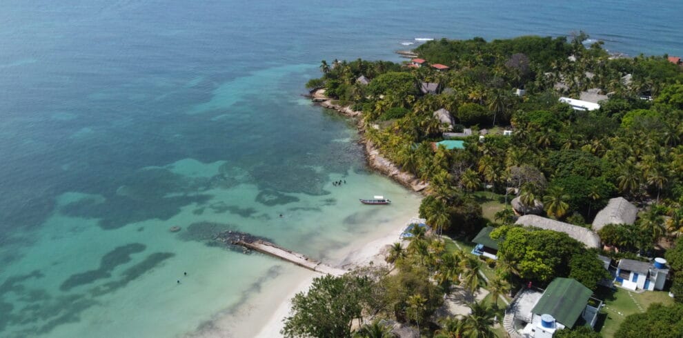 Vista aérea de una costa tropical con aguas cristalinas de color turquesa, una pequeña playa de arena, un barco atracado y una densa vegetación que se extiende hasta la costa.