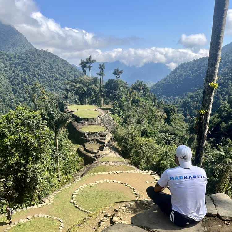 Una persona con gorra y camiseta blanca sentada sobre una roca, proporcionando el telón de fondo perfecto de antiguas terrazas de piedra rodeadas de exuberante vegetación de montaña en un día despejado.