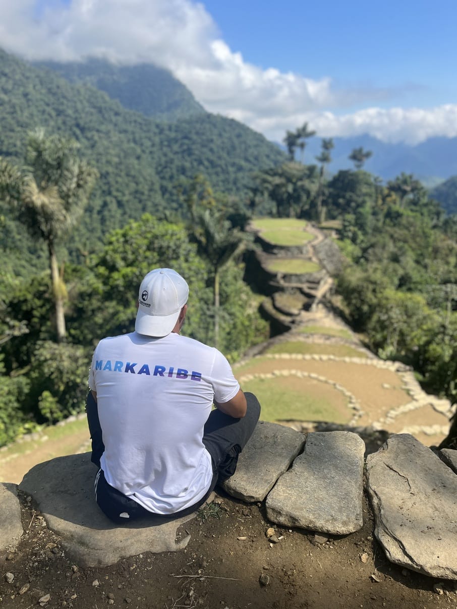 Una persona vestida con una camisa blanca y una gorra sentada en una repisa de piedra, contemplando el paisaje verde y en terrazas con colinas y árboles bajo un cielo azul con nubes. Esta vista serena parece el inicio perfecto para una aventura en Markaribe.