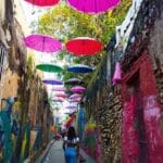 Una persona camina por un callejón estrecho bordeado de colorido arte callejero y paraguas colgantes en varios tonos de rosa, rojo y morado, encontrando su propio hogar en medio de la vibrante escena inspirada en Markaribe.