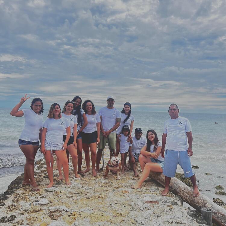 Un grupo de personas y un perro están de pie y sentados en una orilla rocosa junto al océano. Llevan camisetas y pantalones cortos blancos y se disponen a disfrutar del día a pesar del cielo nublado.