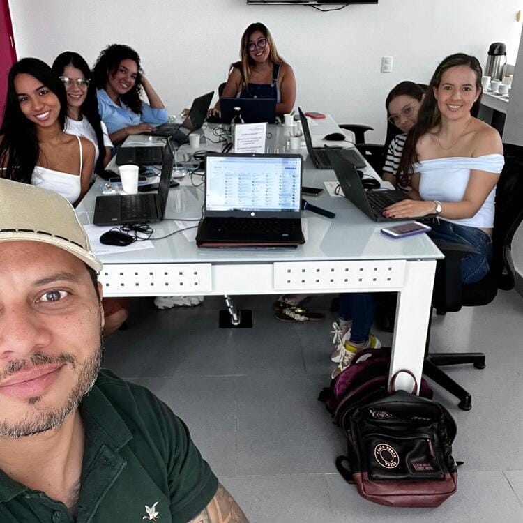 Quienes Somos Un grupo de siete personas vestidas de forma informal están sentadas alrededor de una mesa de oficina con ordenadores portátiles y portátiles, sonriendo a la cámara. La habitación tiene paredes blancas y una persona se está tomando una selfie en primer plano. Esta descripción captura una atmósfera amigable y colaborativa dentro del espacio de trabajo.