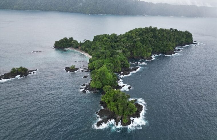 Vista aérea de una pequeña isla boscosa rodeada de costa rocosa y olas, con una masa de tierra más grande visible al fondo bajo un cielo nublado, cerca del Casco Urbano de Nuquí.