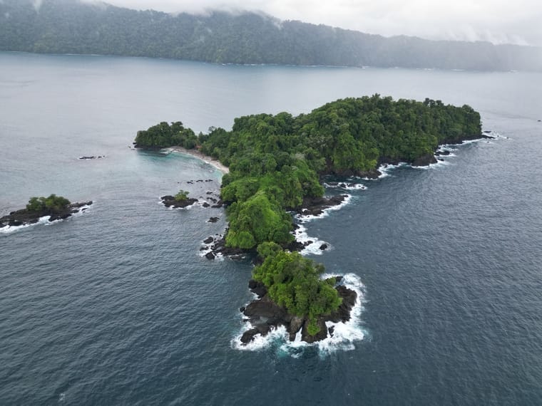 Vista aérea de una pequeña isla boscosa rodeada de costa rocosa y olas, con una masa de tierra más grande visible al fondo bajo un cielo nublado, cerca del Casco Urbano de Nuquí.