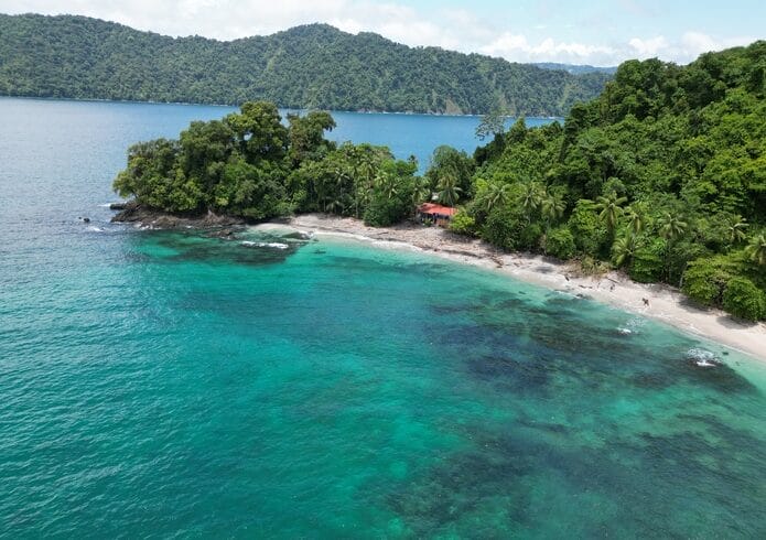 Vista aérea de una playa aislada en Nuquí con aguas cristalinas de color turquesa, rodeada de exuberantes colinas boscosas y un pequeño edificio con techo rojo cerca de la costa. A solo 4 días del concurrido Casco Urbano, es una escapada perfecta.